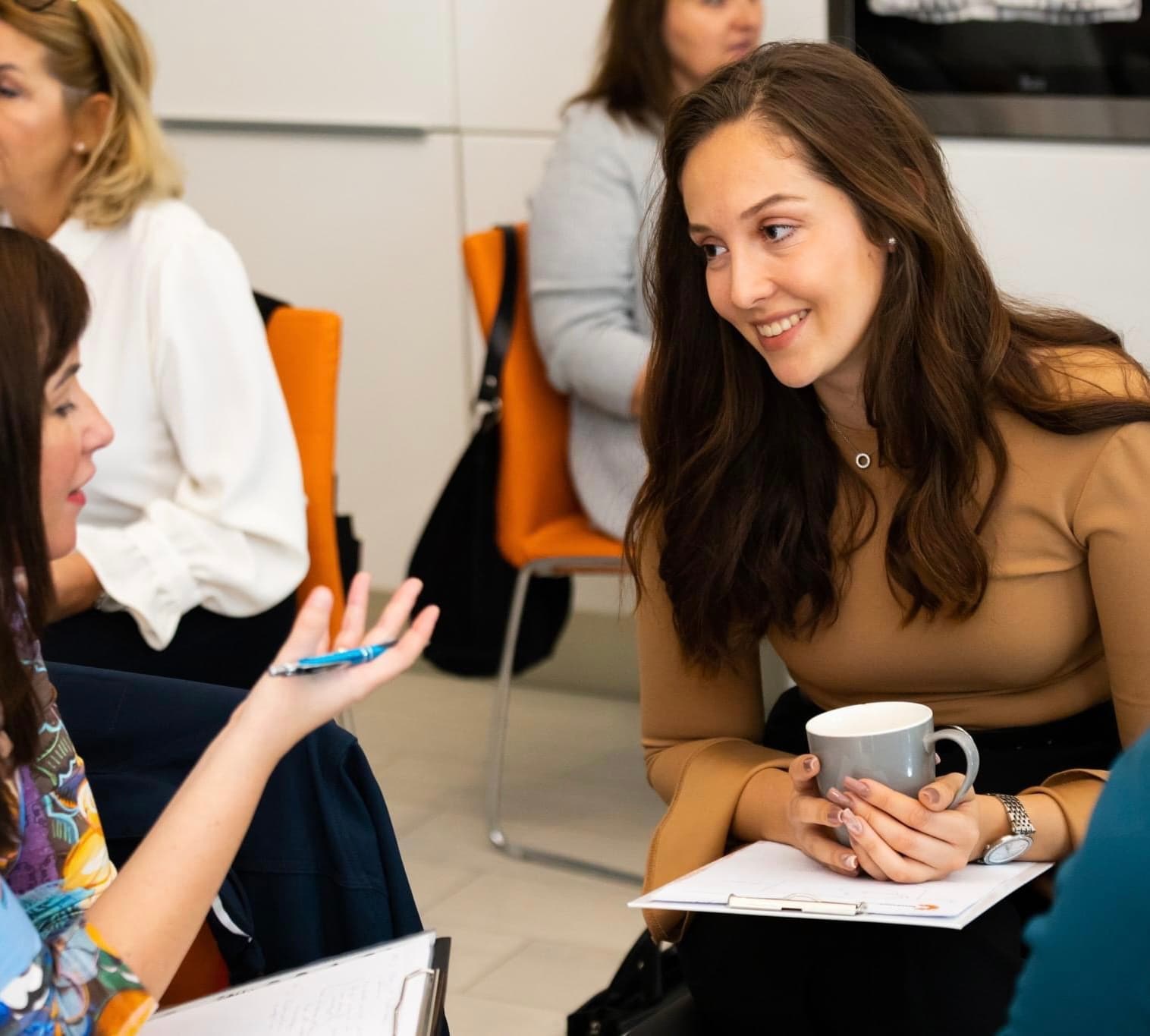 Lächelnde Frau mit braunen Haaren hält eine Tasse und hört bei einem Workshop aufmerksam zu.
