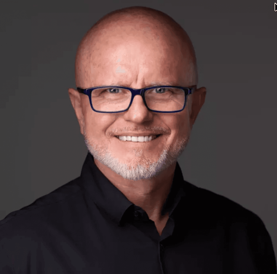 Porträt eines lächelnden glatzköpfigen Mannes mit blauer Brille, grauem Bart und schwarzem Hemd.